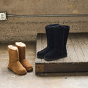 Two pairs of mid-calf suede boots, one in tan and one in black, displayed on a rustic wooden platform against a concrete wall.