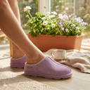 A lifestyle shot of a woman’s feet wearing lilac suede Bearpaw clogs with a woven chevron-patterned trim. She is sitting on a light wood floor in a sunlit room, next to a terracotta planter filled with white and purple spring flowers. A ceramic coffee mug and a soft linen cloth rest nearby, creating a bright and cozy spring atmosphere.