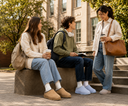 Three young adults in casual campus attire converse on outdoor stone steps in front of a university building. One person sits on a bench wearing beige slippers, while the others wear blue slippers.